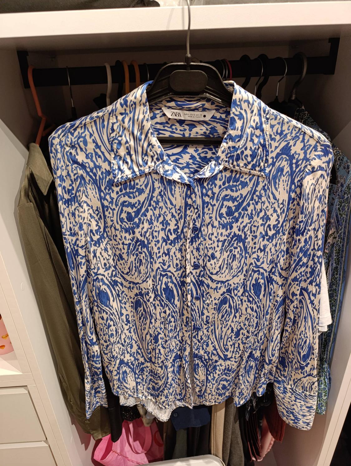 White shirt with blue print hanging in a wardrobe. Label: ZARA EUR S USA S MEX 26 MADE IN TURKEY.