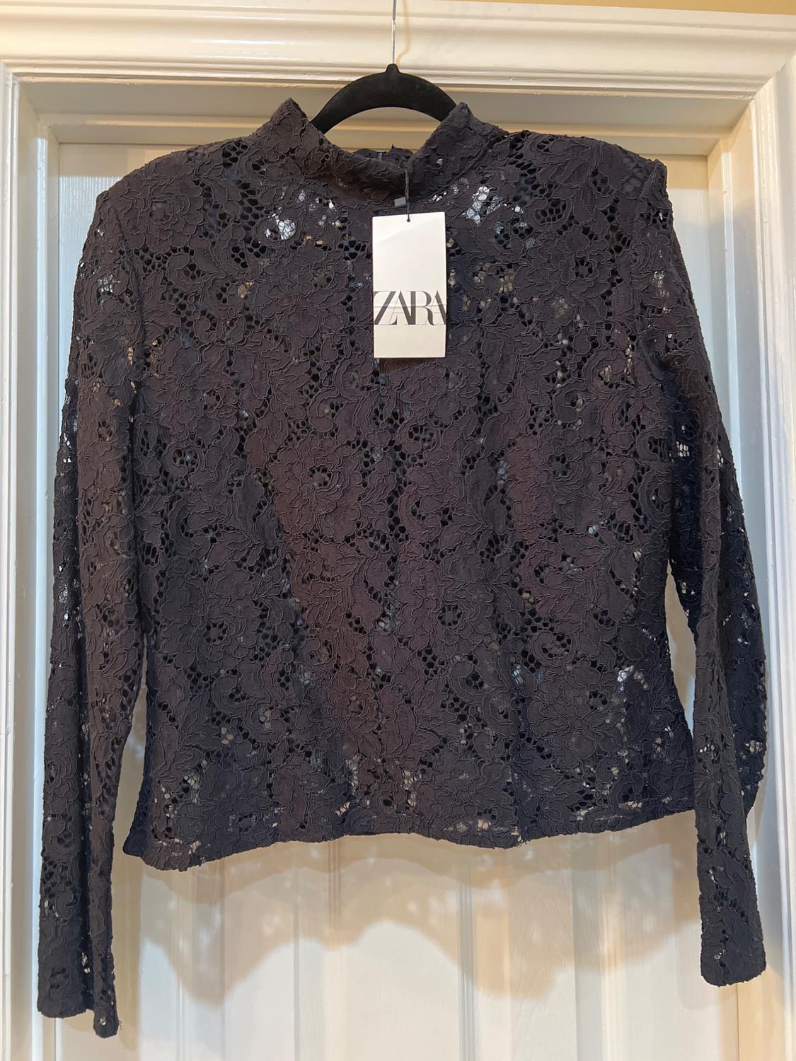 Black lace blouse with long sleeves and a high neck, hanging on a hanger. Visible label with the text "ZARA".