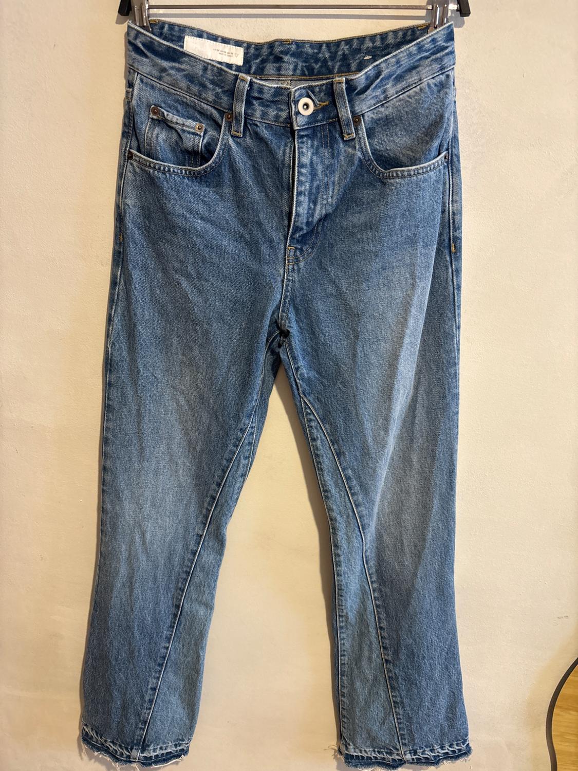 Worn blue jeans hanging on a hanger against a beige background. They have a classic five-pocket design and a frayed hem.