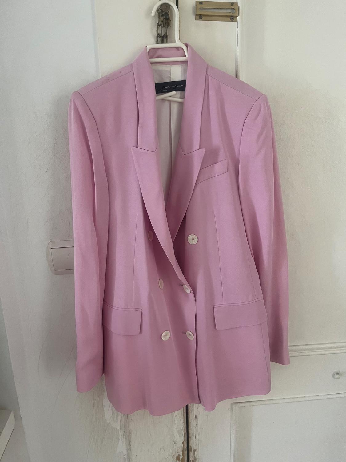 Light pink blazer hanging on a white hanger over a door. Visible label: "ZARA WOMAN". White buttons and front pockets.