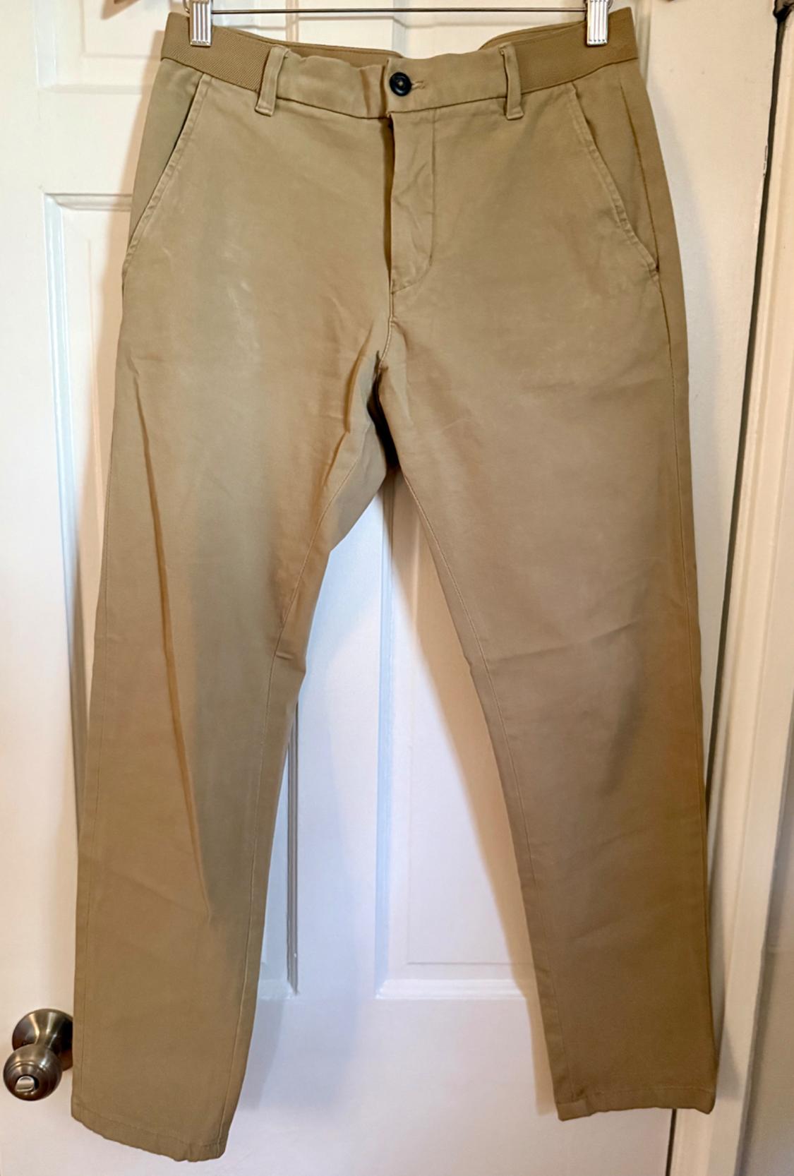 Beige chinos pants hanging on a white door from a hanger. They feature a button front and an elastic waistband.