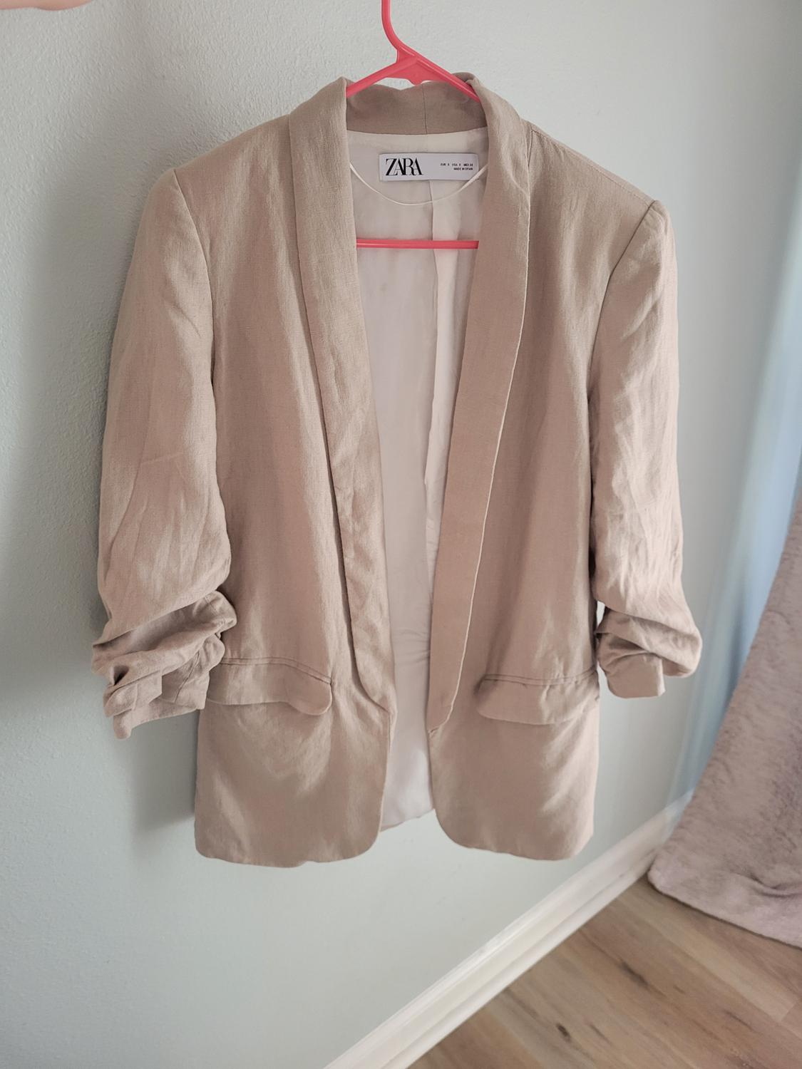 Beige blazer hanging on a pink hanger, with rolled-up sleeves and a visible tag inside. Light wall background and wooden floor.