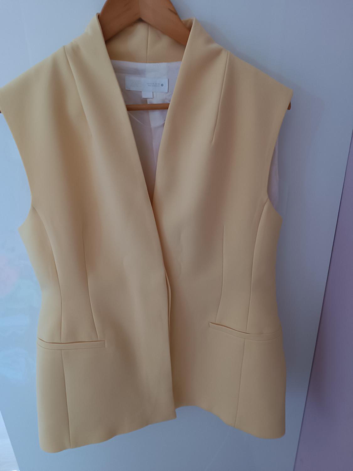Sleeveless beige waistcoat hanging on a wooden coat hanger, with a white background and visible stitching details.