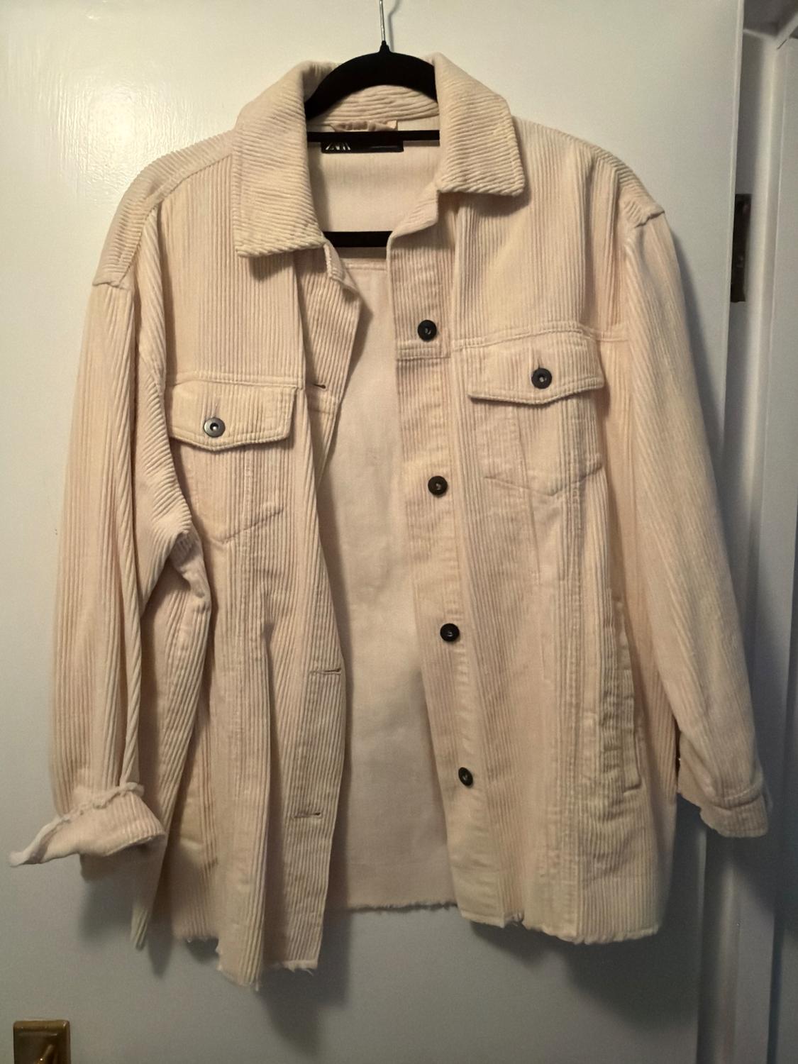 Beige corduroy jacket hanging on a black hanger over a white door, with front pockets and dark buttons.
