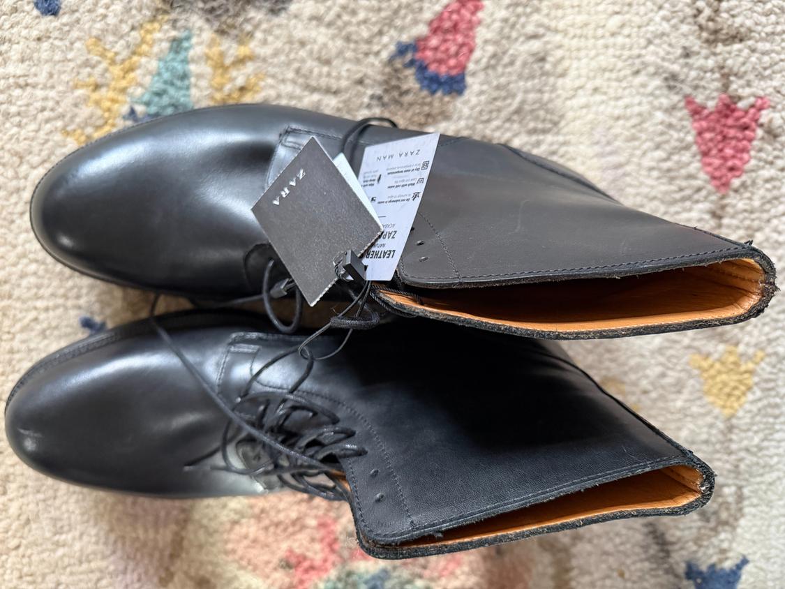 Black leather boots with a brand label on a colourful rug. Top-down view showing design details and laces.