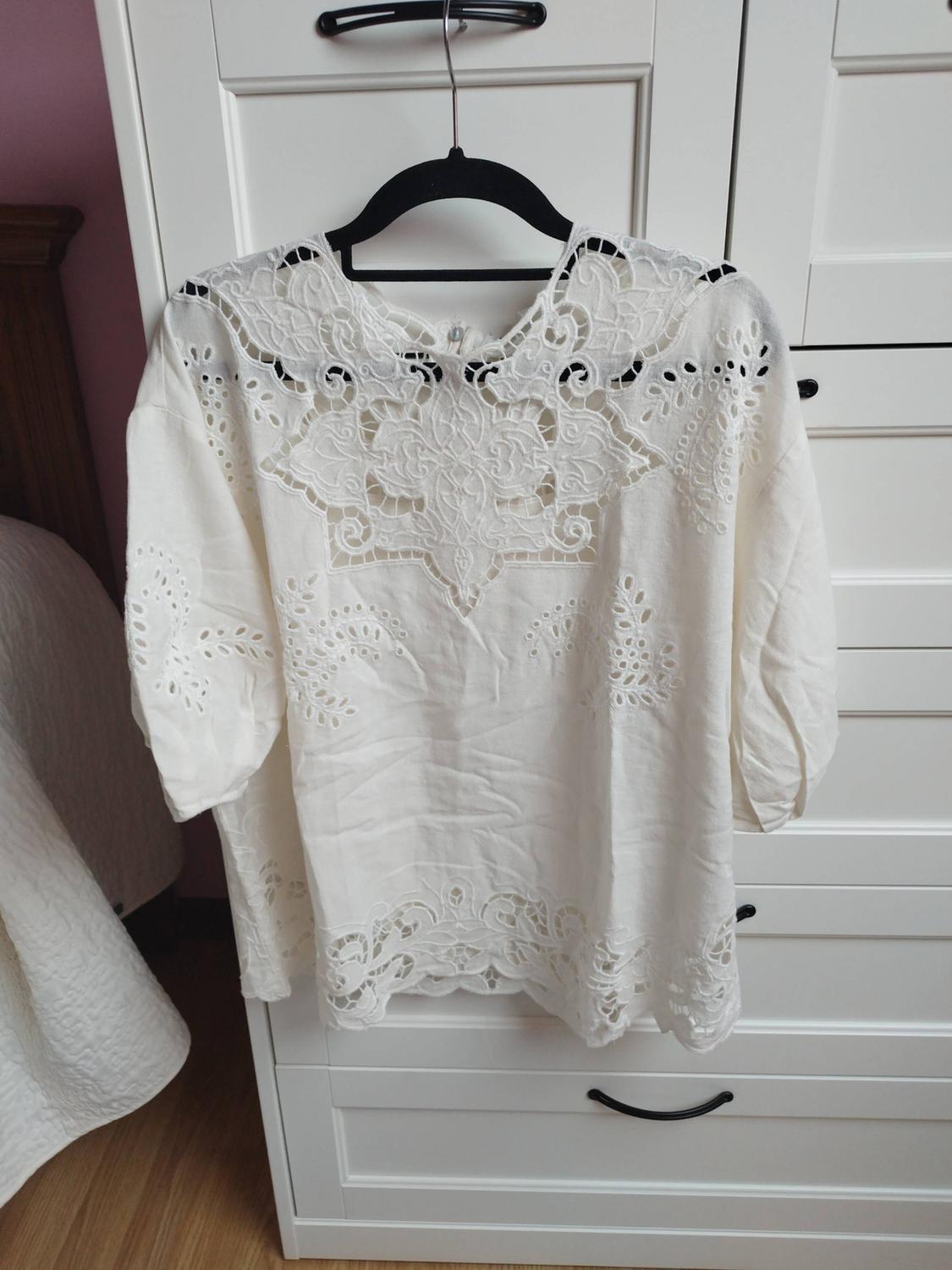 White short-sleeved blouse with openwork embroidery, hanging in a white wardrobe. Relaxed bedroom setting with a bed and wooden floor.