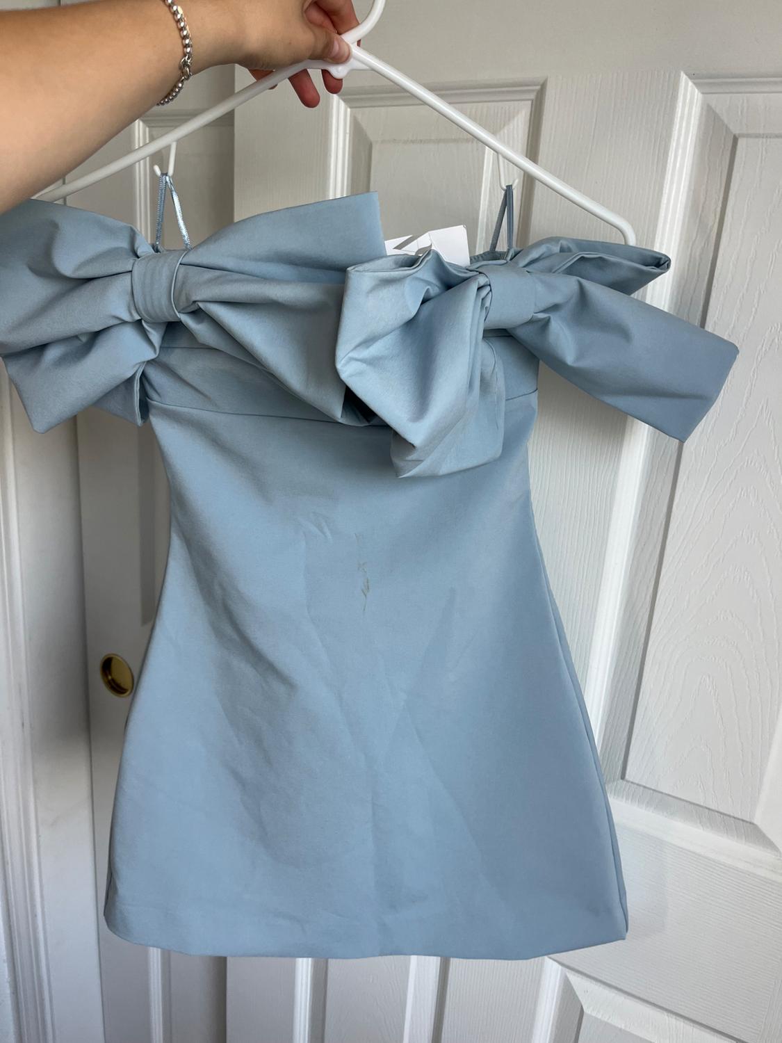 Light blue dress with large bows on the shoulders, hanging on a white hanger. White door background. Hand holding the hanger.