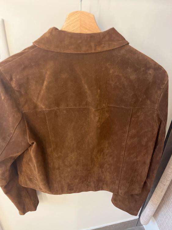 Brown suede jacket hanging on a wooden hanger, seen from behind, against a light background in a store.