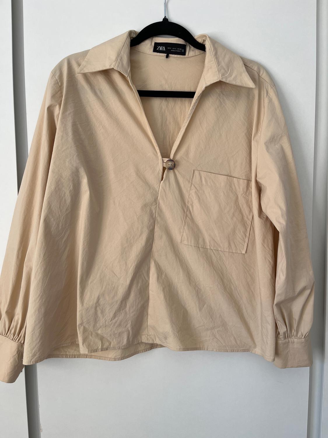 Long-sleeved beige shirt with a visible button and chest pocket, hanging on a black hanger against a white background.