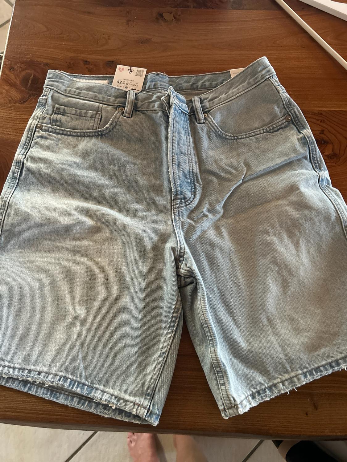 Light blue denim shorts on a wooden table. They have frayed hems and a label bearing the text 42 32 32 32 48 42.