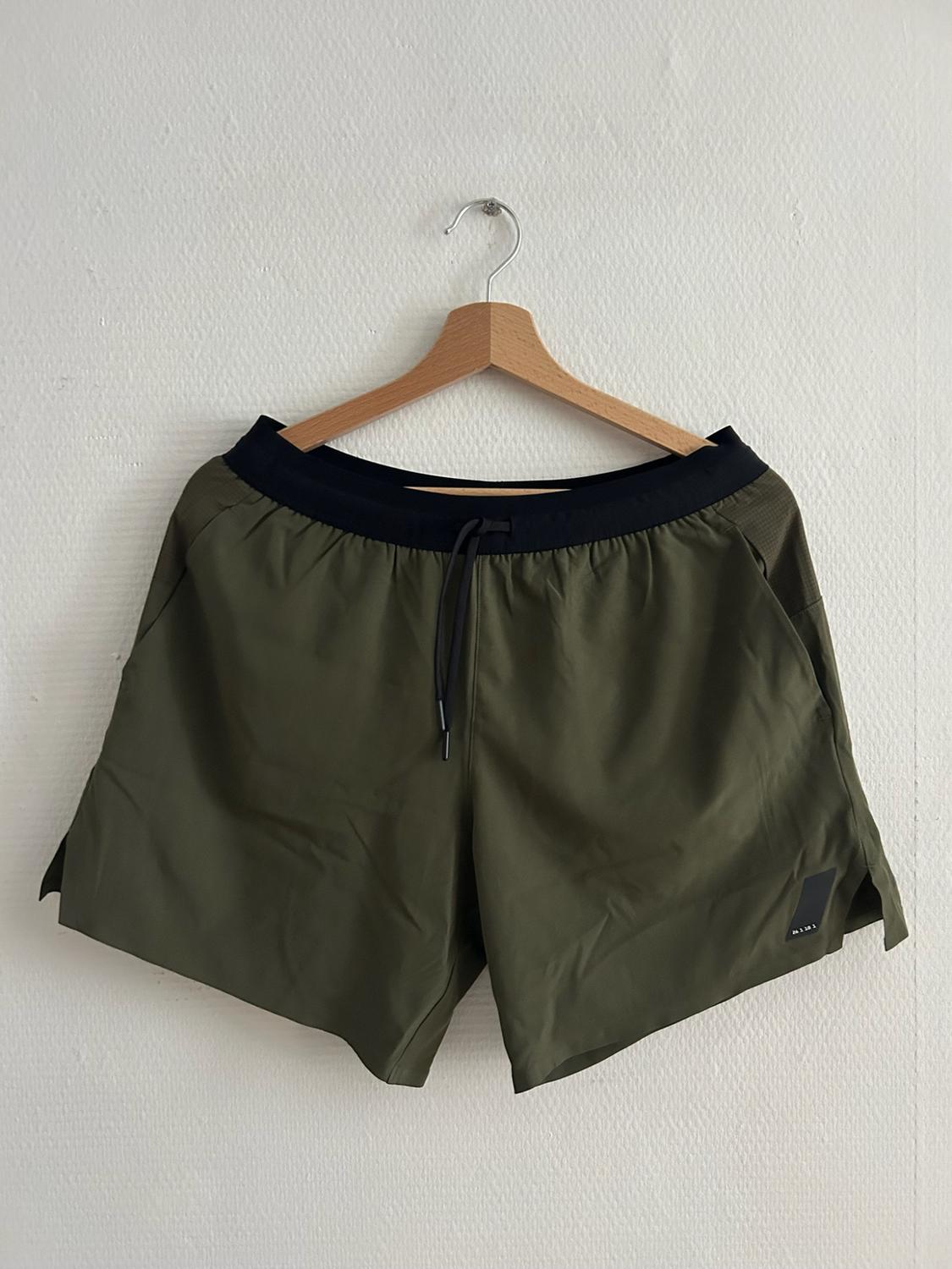 Olive green sports shorts with a black elasticated waistband and drawstring, hanging on a wooden hanger against a white wall.
