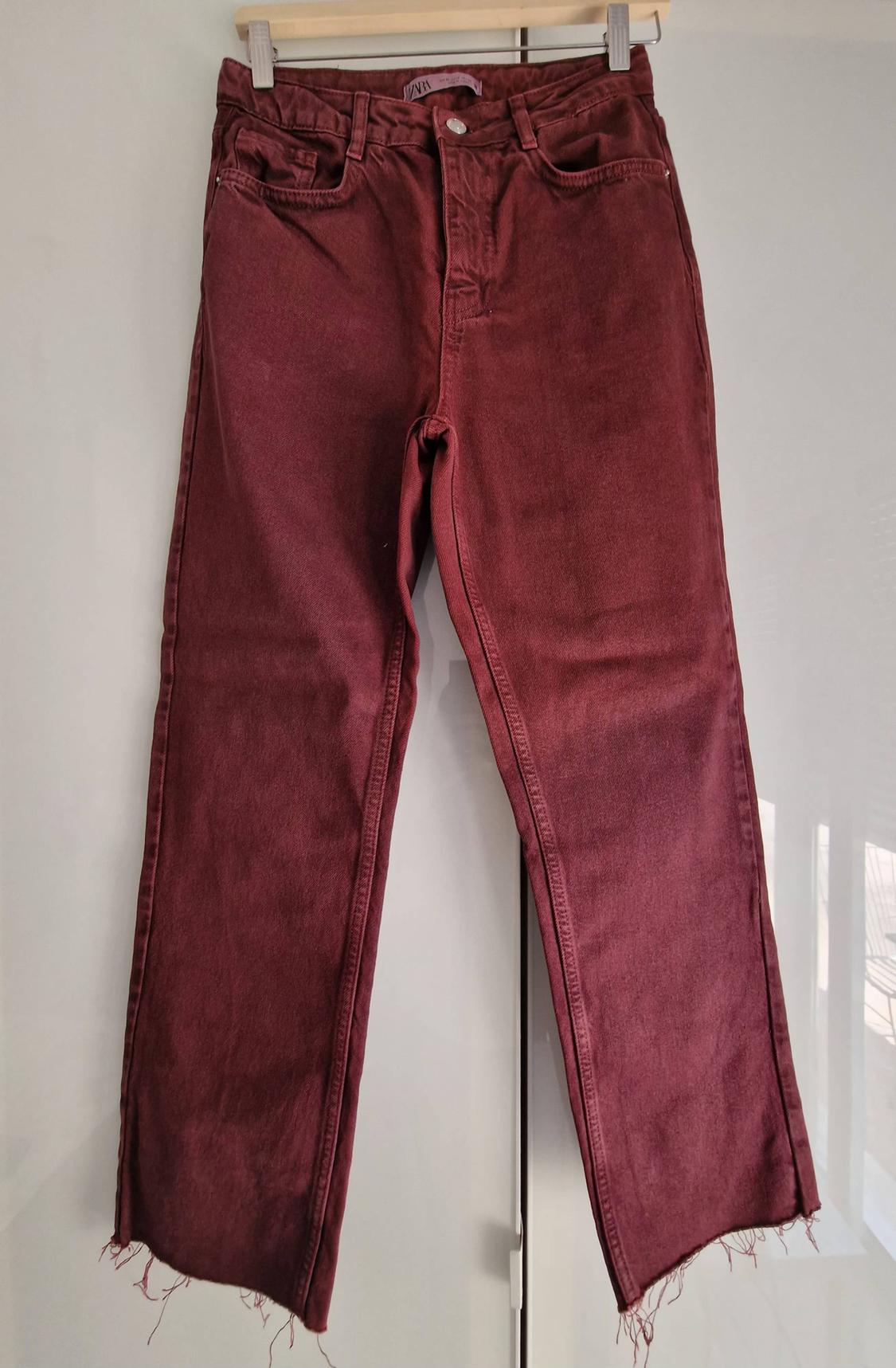 Burgundy jeans with a frayed hem hanging on a hanger in front of a white wardrobe. ZARA label visible.