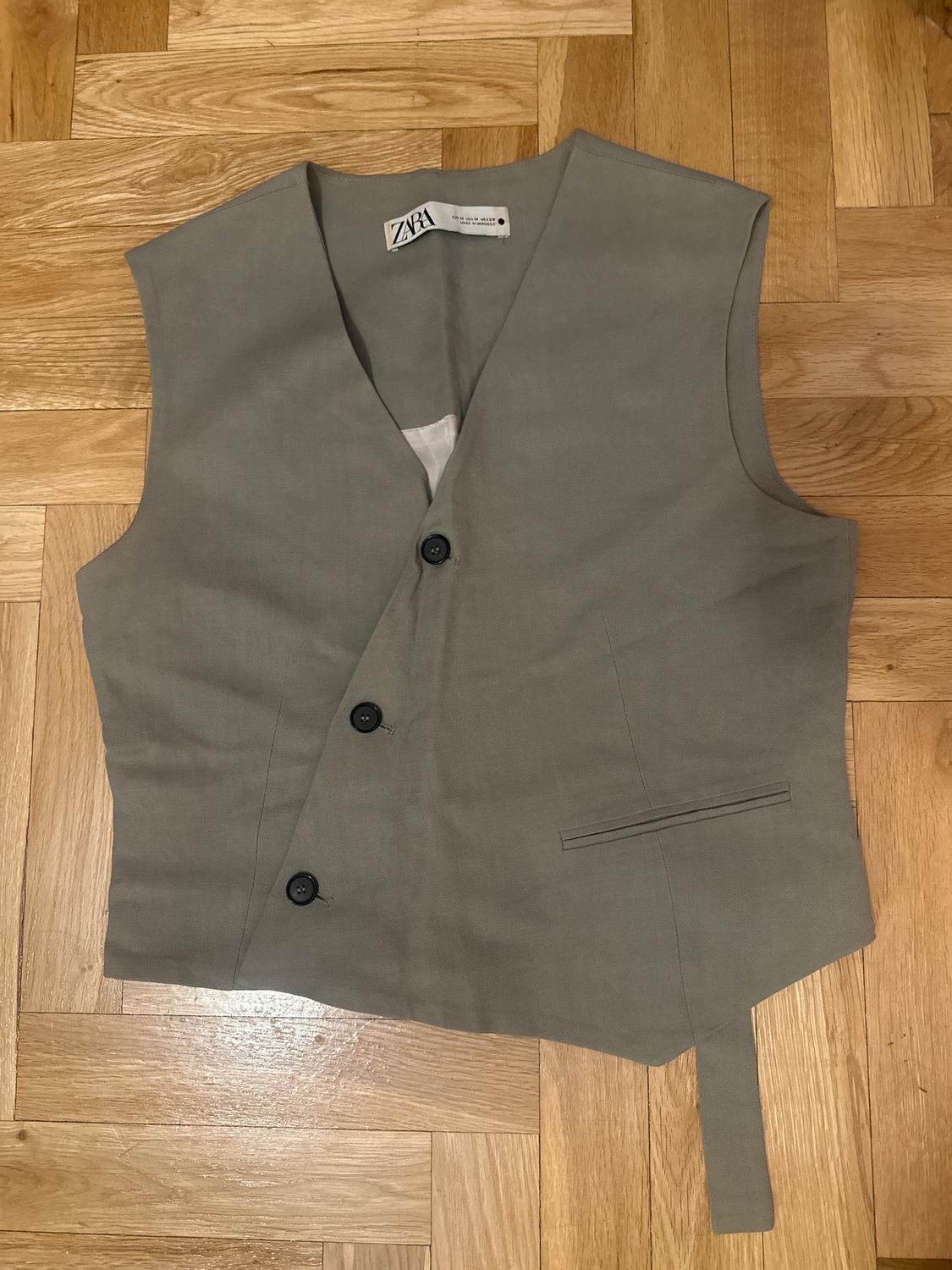 Sleeveless grey waistcoat with three black buttons on a wooden floor. Visible label with text: "ZARA".