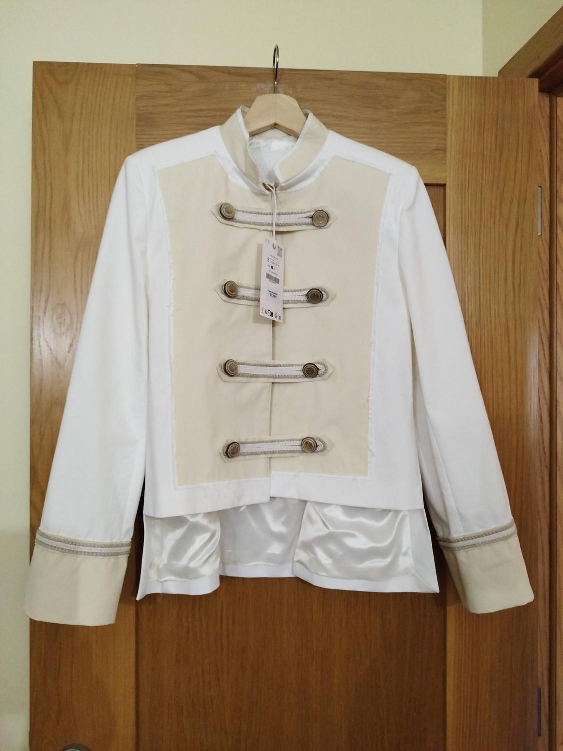 White and beige military-style jacket with decorative front buttons and laces, hanging on a hanger over a wooden door.