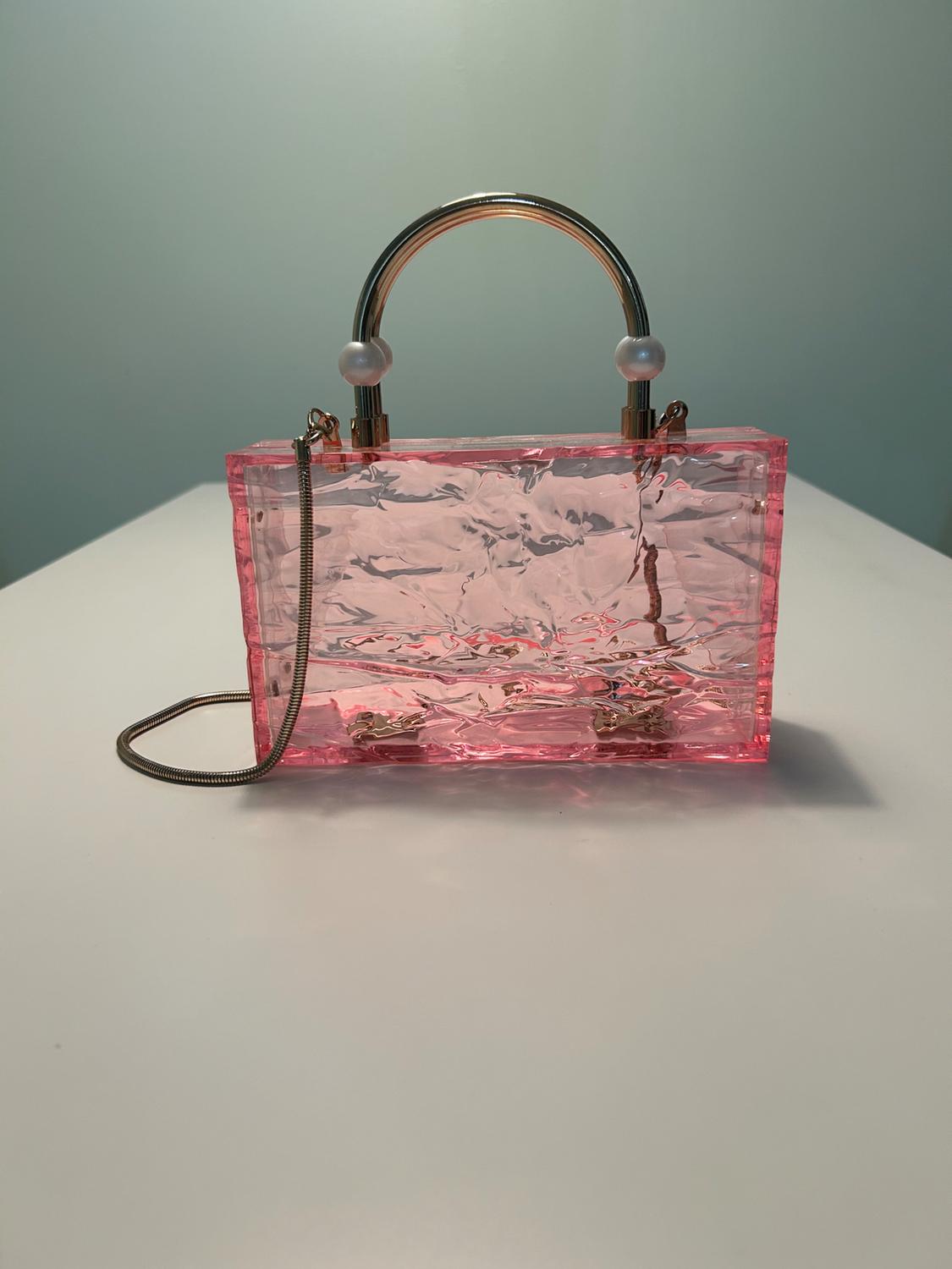 Transparent pink rectangular bag with a crinkled texture on a white surface. It features a metal handle with pearls and a thin chain.