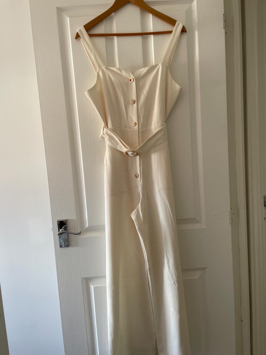 Cream-coloured long jumpsuit with front buttons, pockets and a belt, hanging on a wooden hanger on a white door.