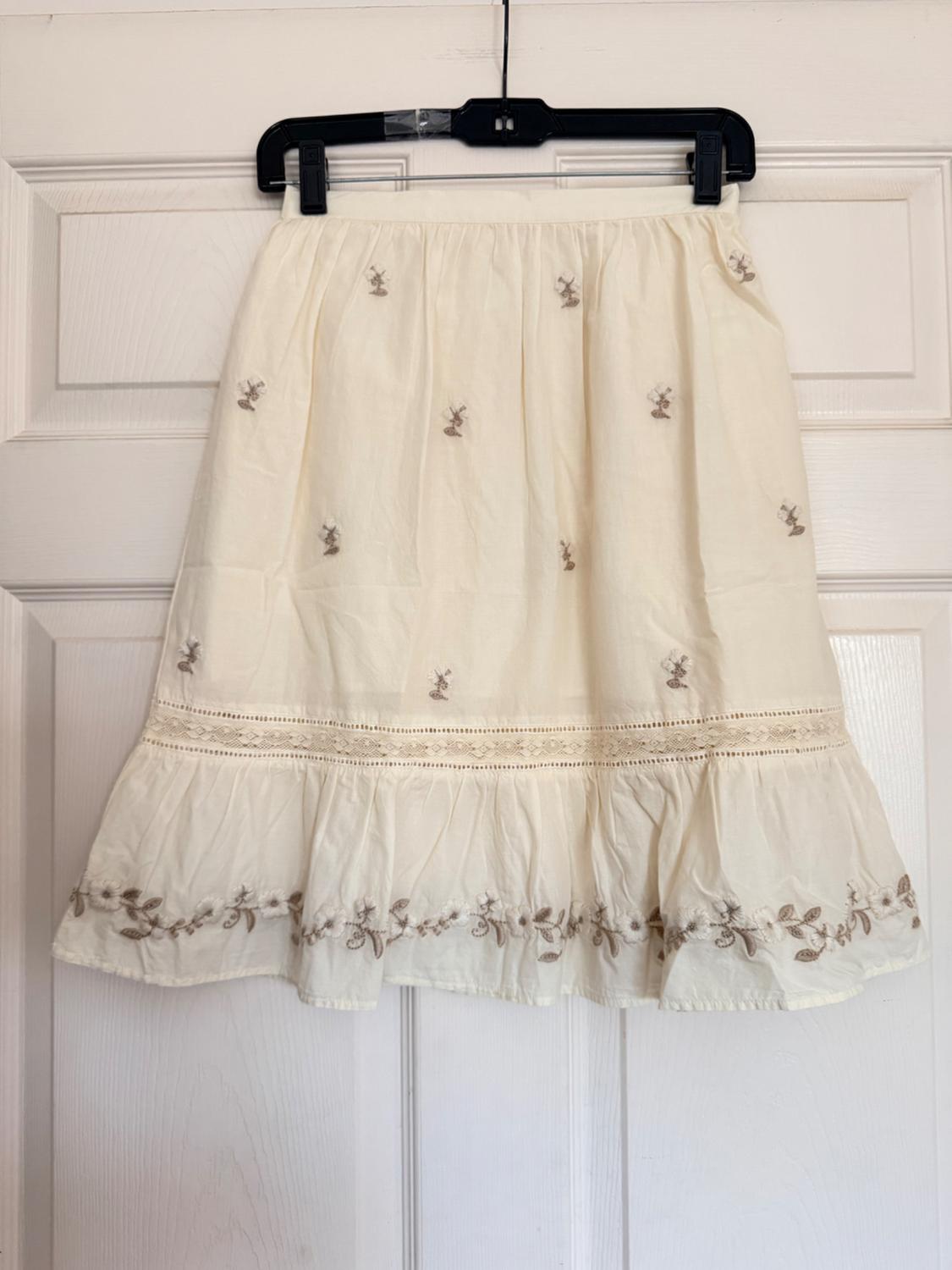 Cream-colored skirt hanging on a white door. It features small floral embroidery, a lace trim, and an embroidered ruffled hem.