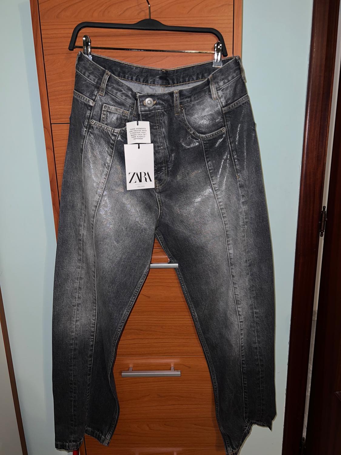 Worn grey denim jeans hanging on a black hanger with a Zara Studio tag, light wood background.