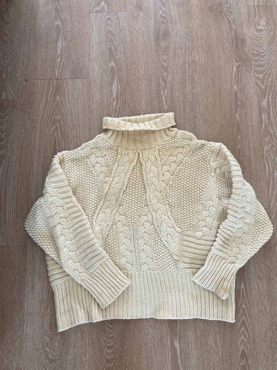 Cream-colored chunky knit sweater with a high neck and cable-knit design, on a light wooden floor.