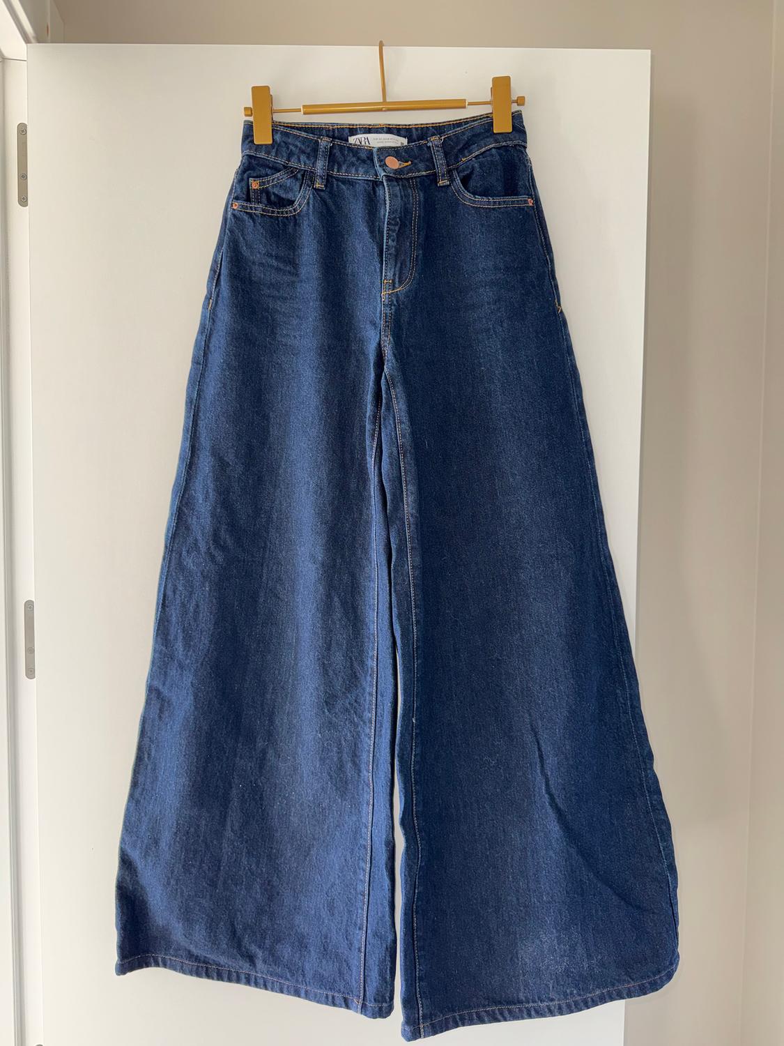 Wide-leg jeans hanging on a wooden hanger over a white door. Label visible on the waistband.