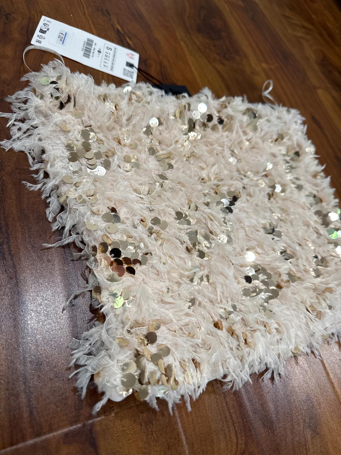Beige fabric with gold sequins and a furry texture on a wooden floor. Visible label with size S and barcode.