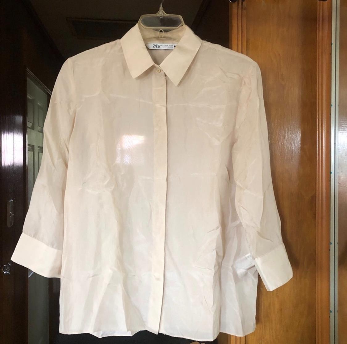 Long-sleeved beige shirt hanging on a hanger, with a wooden door background. Visible tag: "ZARA".