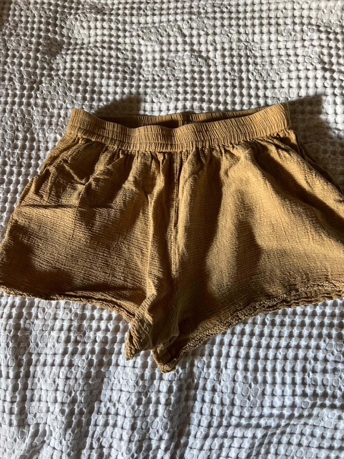 Mustard-coloured textured shorts with an elasticated waist, resting on a white textured bedspread with natural light shadows.