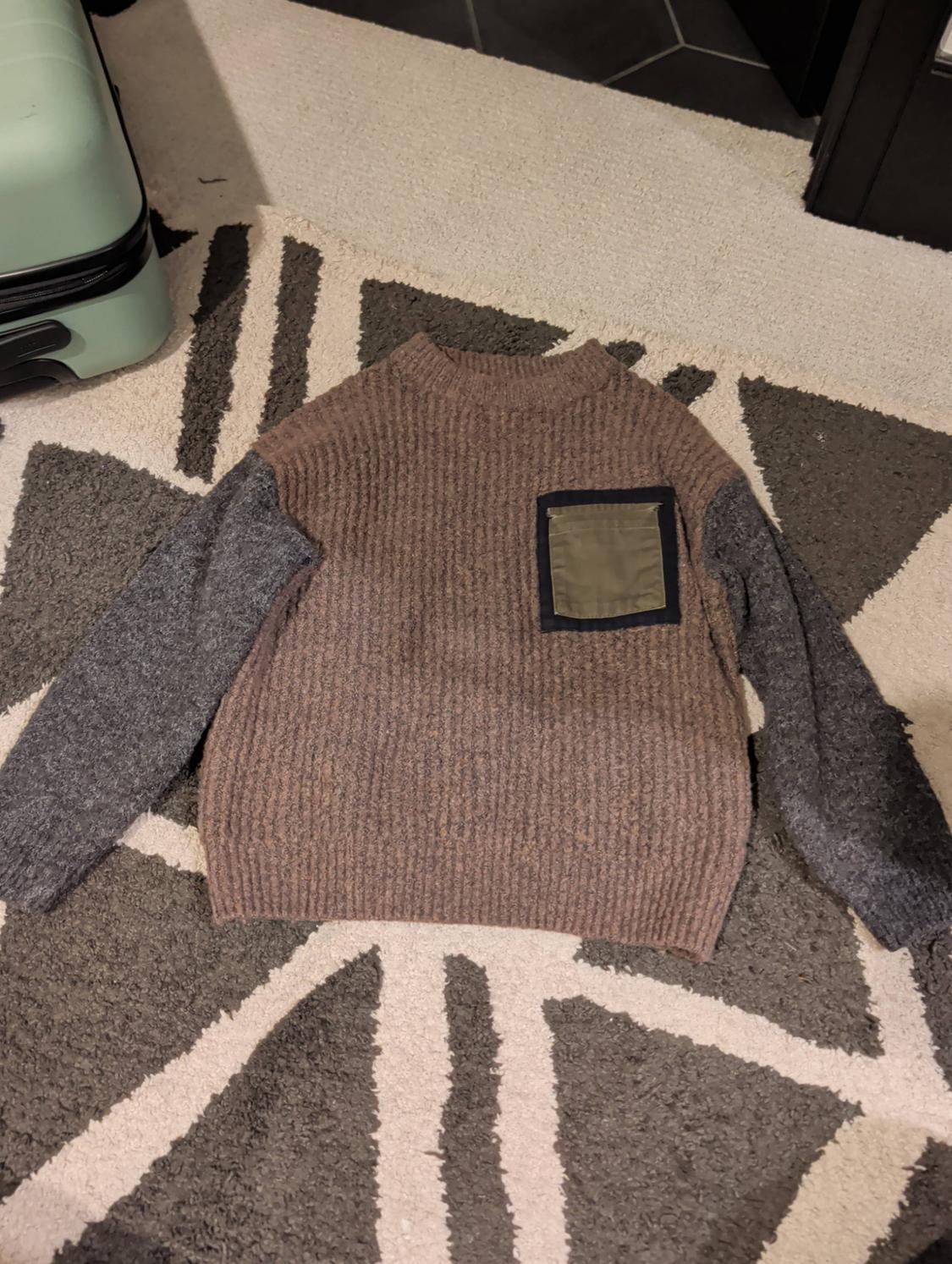 Knitted jumper with a brown body, grey sleeves and a green pocket, on a grey and white geometric rug.