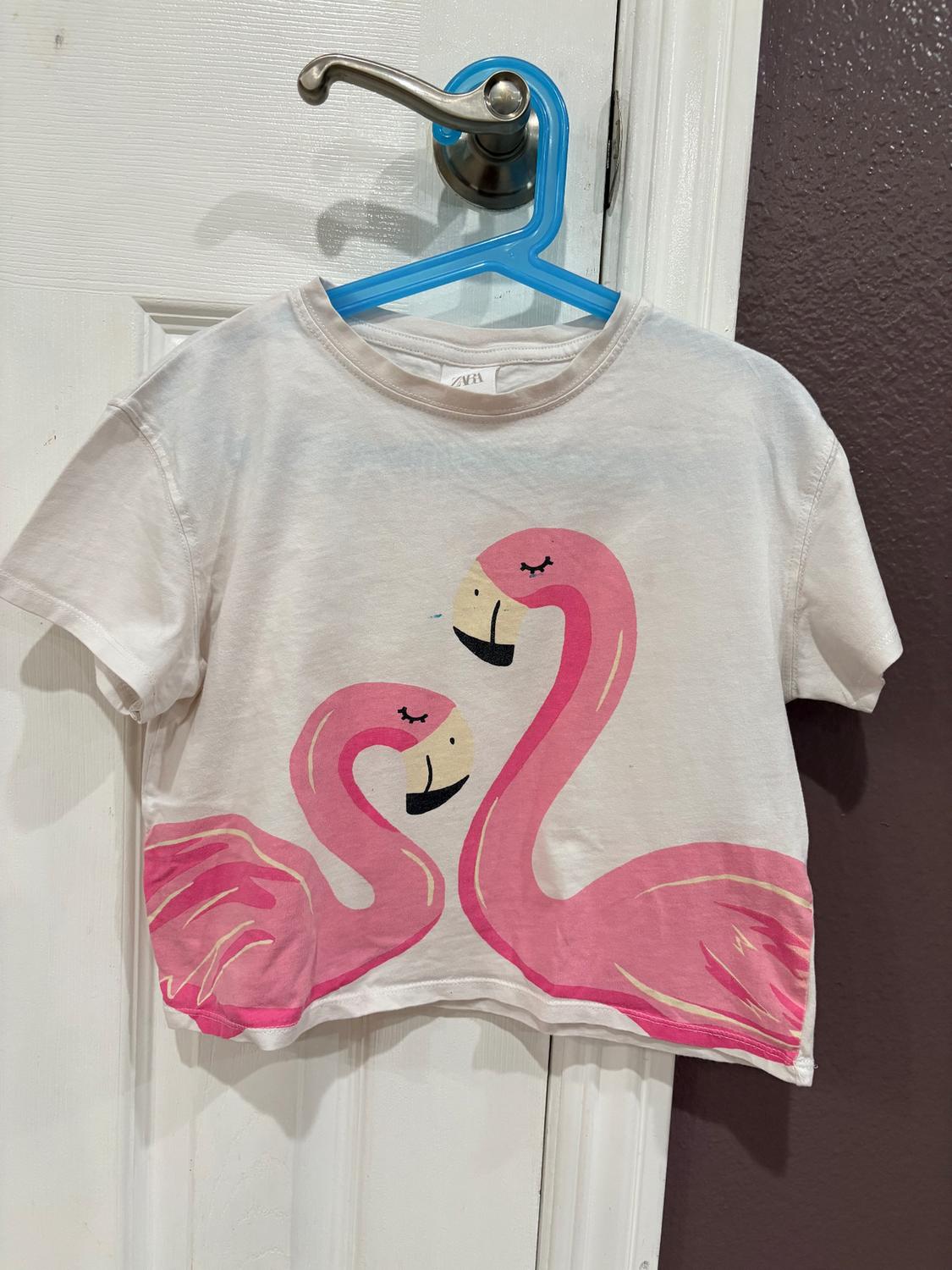 White t-shirt with a print of two pink flamingos facing each other. It is hanging on a blue hanger over a white door.