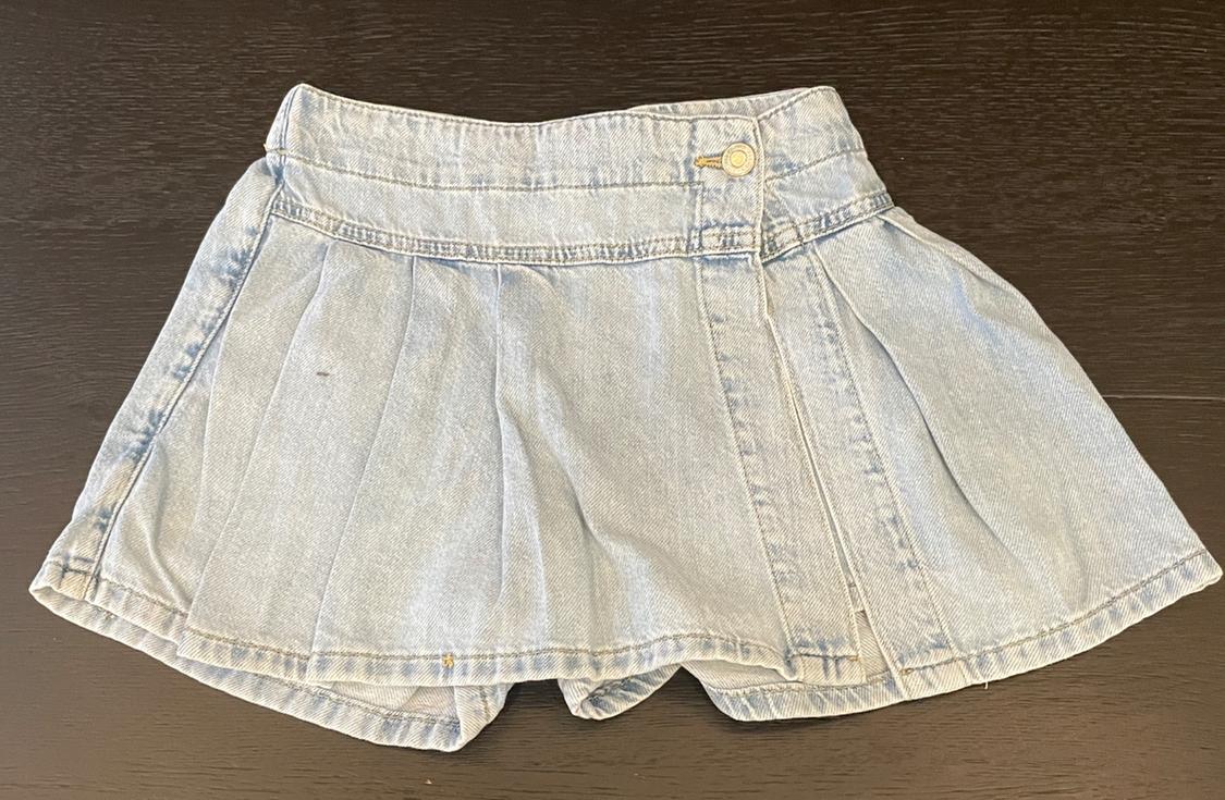 Light blue pleated denim skort with asymmetric button fastening, placed on a dark wooden surface.