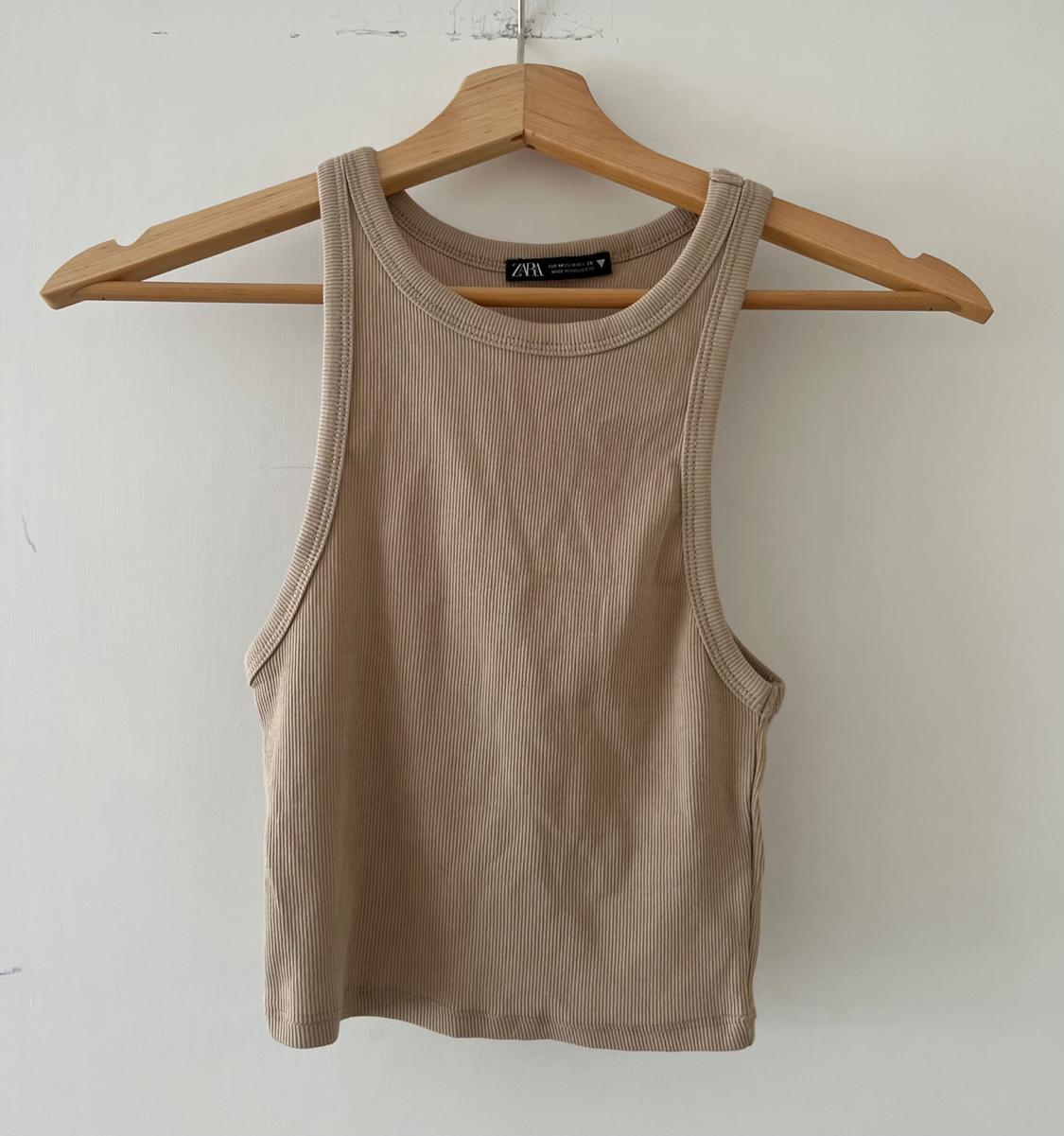 Beige ribbed sleeveless crop top hanging on a wooden hanger against a white wall. Label with ZARA text. Casual style.