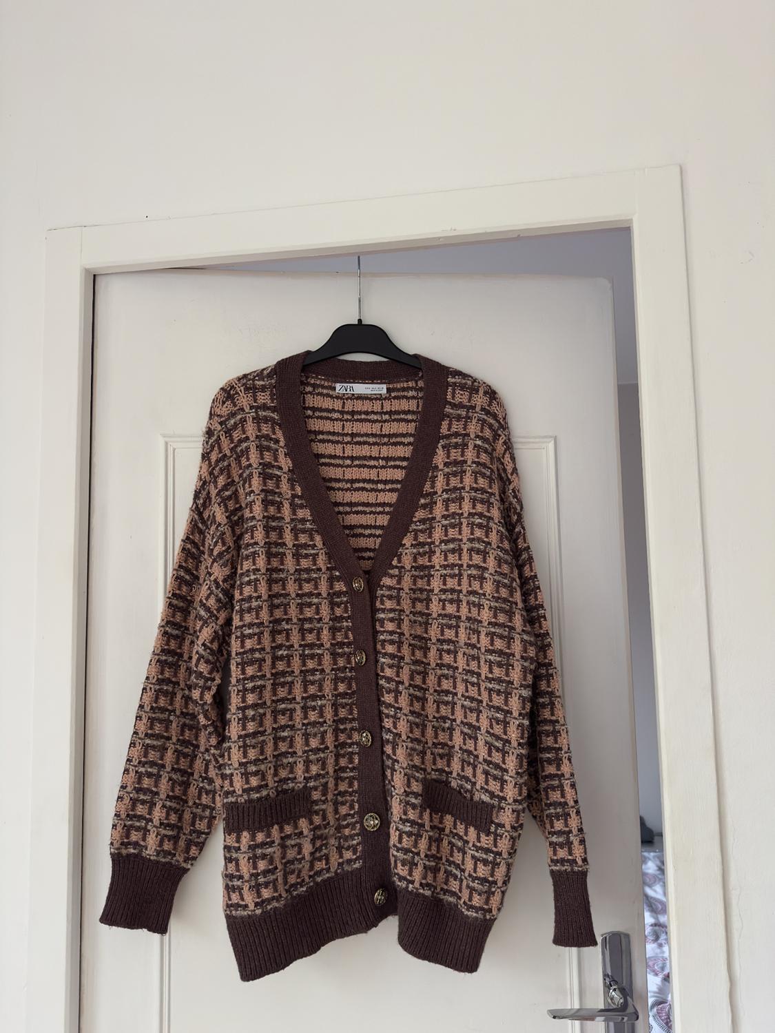 Brown cardigan with a geometric print, hanging on a black hanger over a white door. Visible label: ZARA.