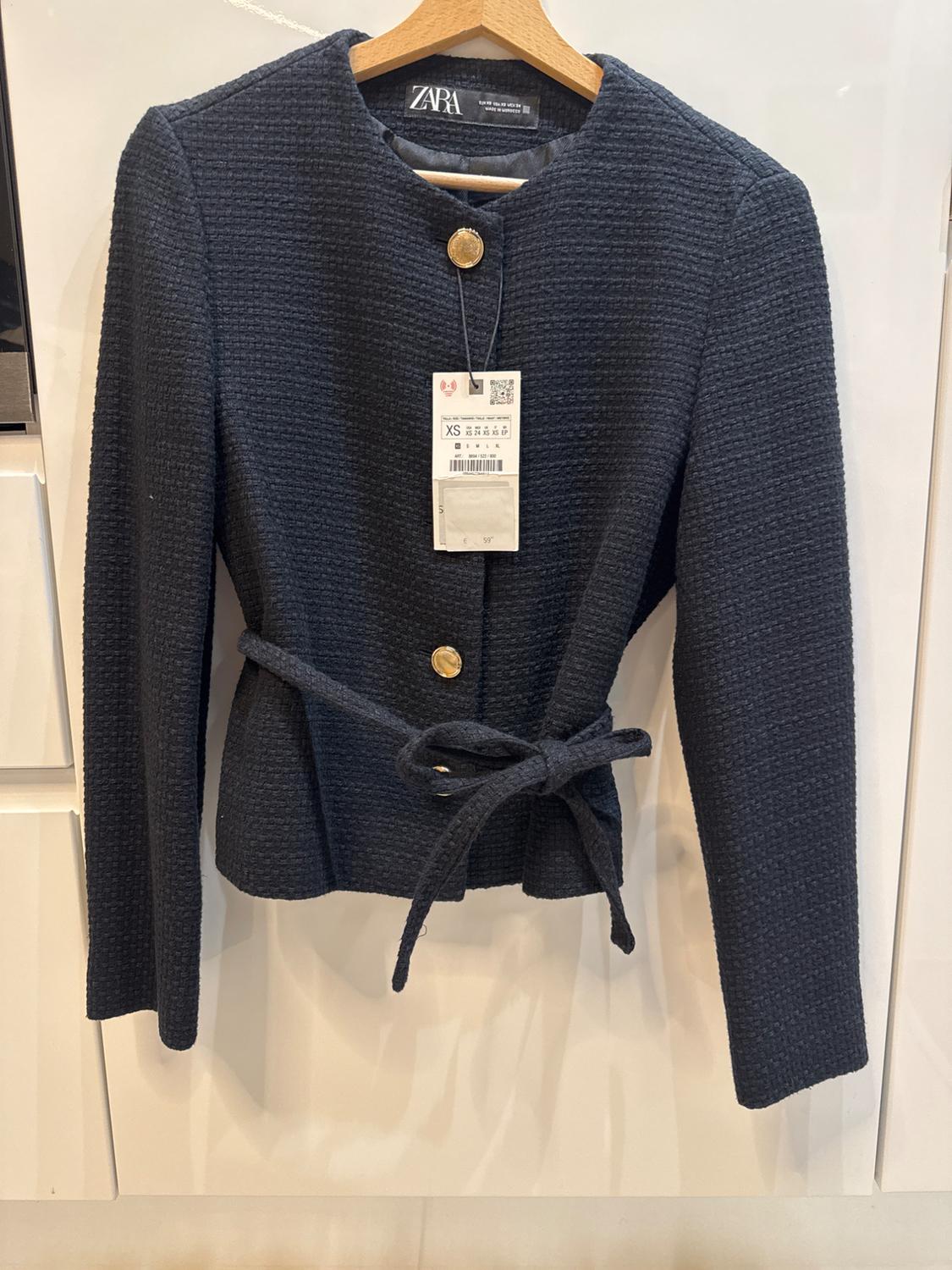Black Zara jacket with texture, gold buttons, and belt, hanging on a wooden hanger. Visible label with size XS.