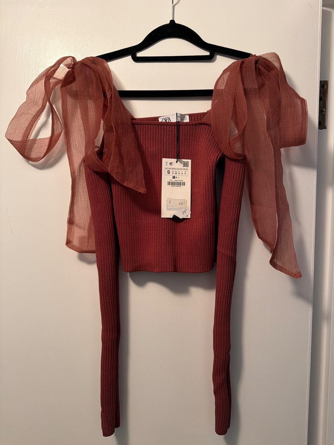 Red knitted long-sleeved top with chiffon ties on the shoulders, hanging on a black hanger. Visible price tag.