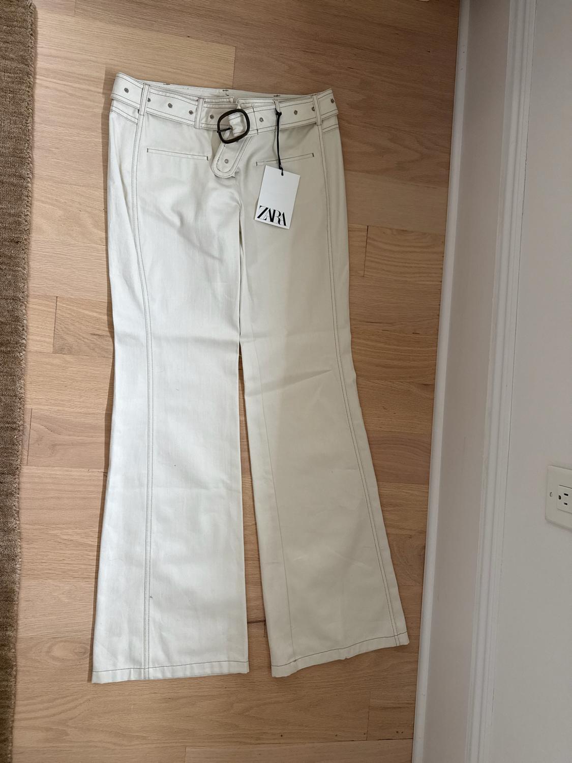 Cream-colored flared trousers with a belt and front seams, on a wooden floor. Visible label with the text ZARA.
