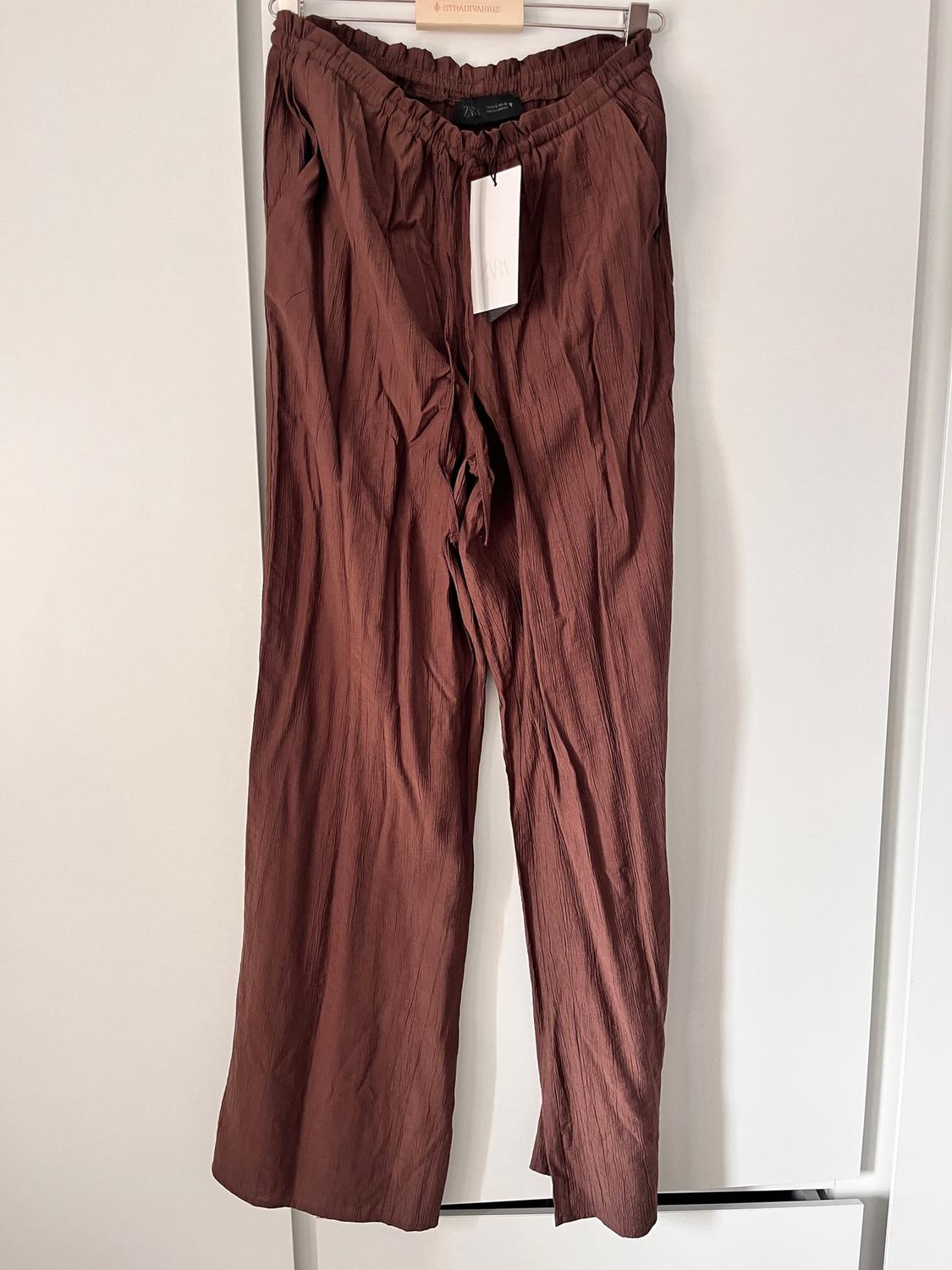 Creased brown trousers hanging on a white hanger, with a white tag visible at the top. White background.