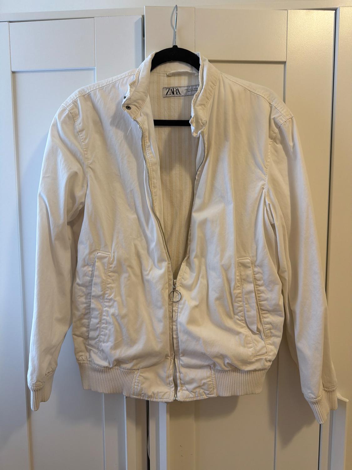Casual white zip-up jacket with pockets, hanging on a black hanger over a white door. Label with the text ZARA.