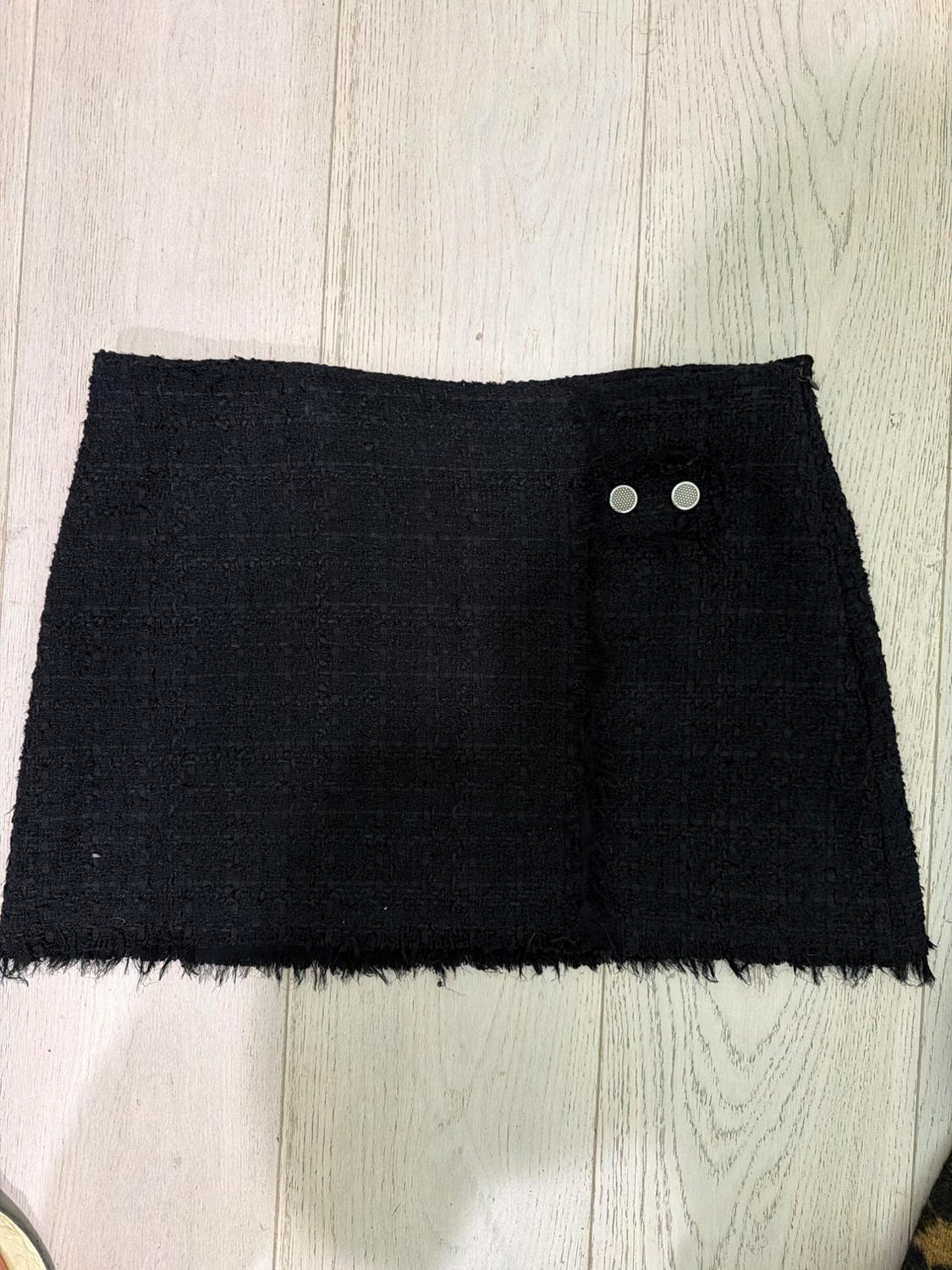 Black textured fabric mini skirt with a frayed hem and two silver-toned buttons, on a light wood floor.