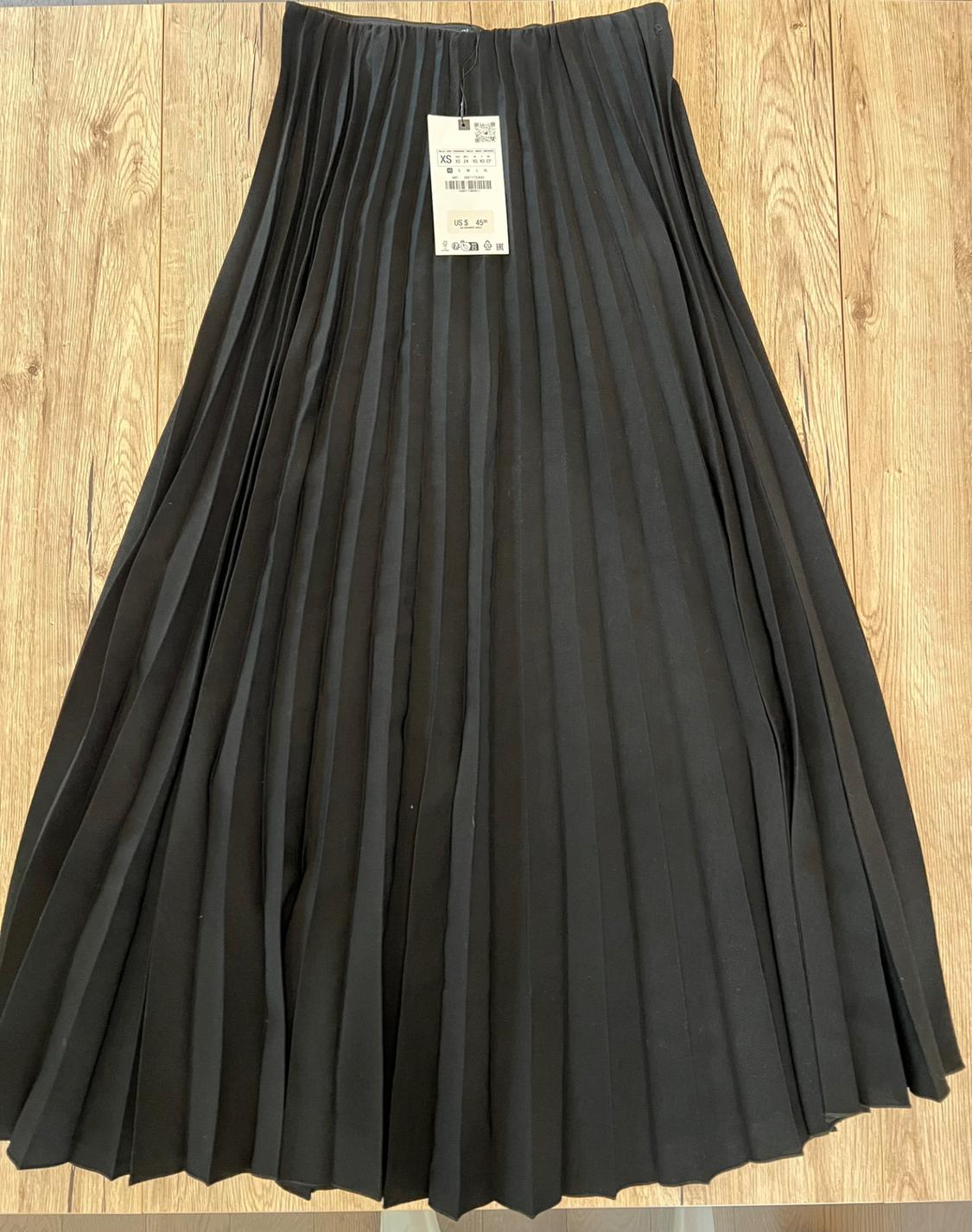 Black pleated skirt laid out on a wooden floor. There's a tag on the waistband with the text XS and US $45.
