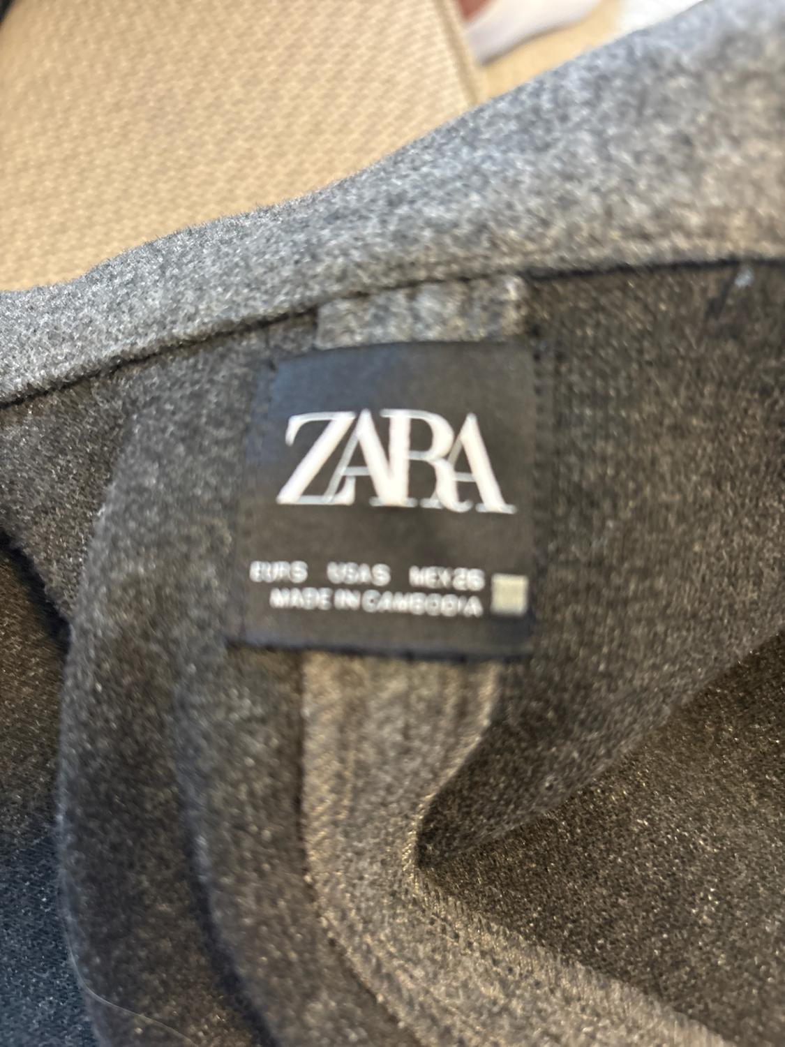 Close-up of a black label on a dark grey garment with the text: ZARA EUR S USA S MEX 26 MADE IN CAMBODIA.