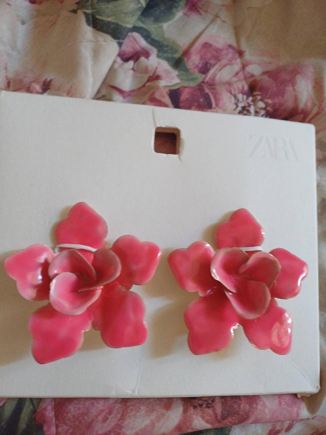 Two pink flower-shaped earrings on white card with the text ZARA, against a floral print fabric background.