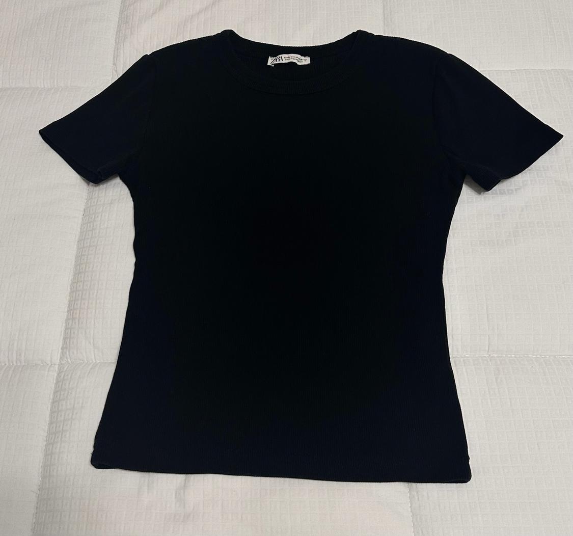 Black short-sleeve, crew-neck T-shirt on a white quilted surface. Label with ZARA text visible at the neckline.