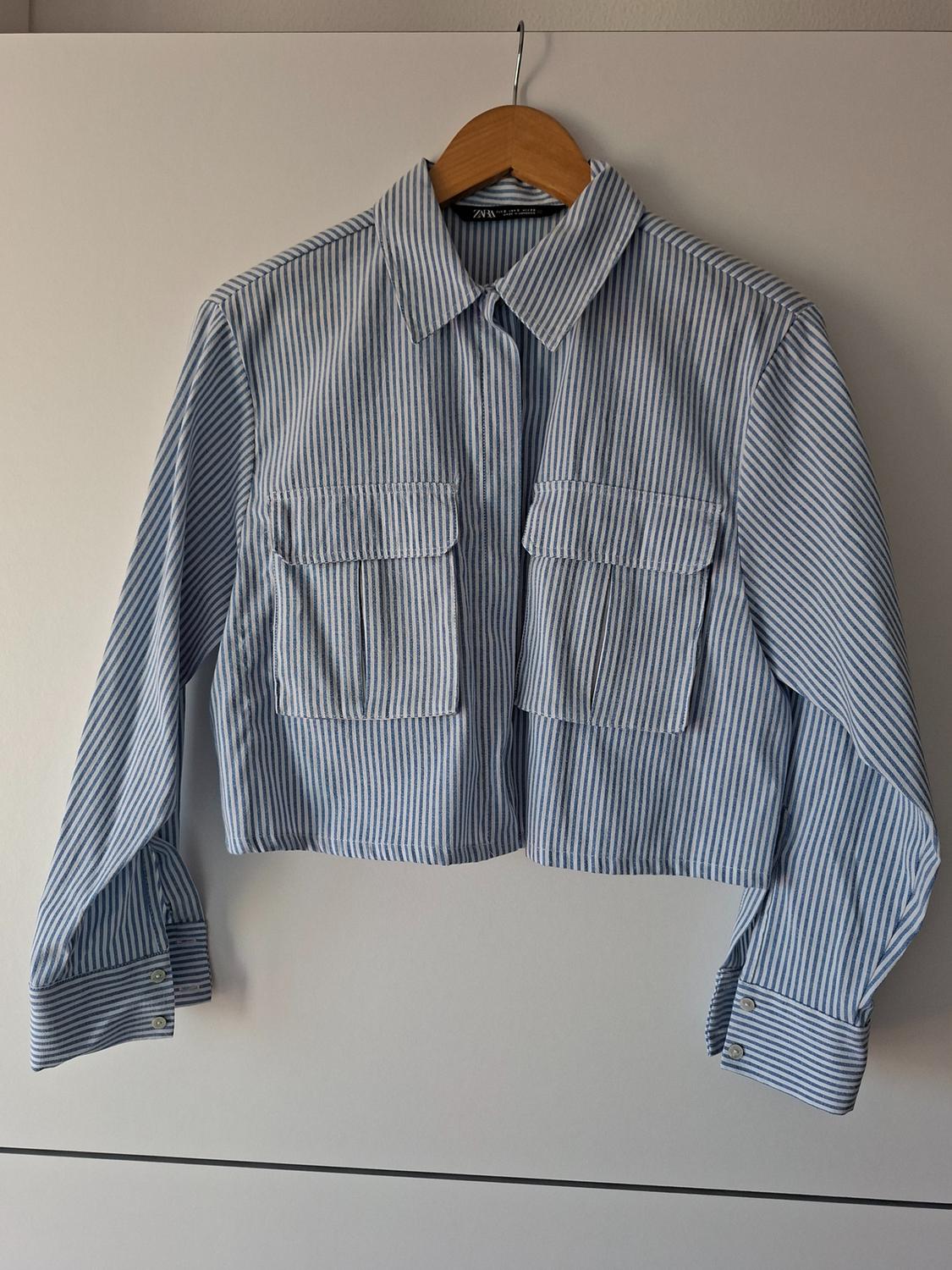 Cropped long-sleeved shirt with blue and white stripes, collar and two chest pockets, hanging on a hanger. ZARA text.