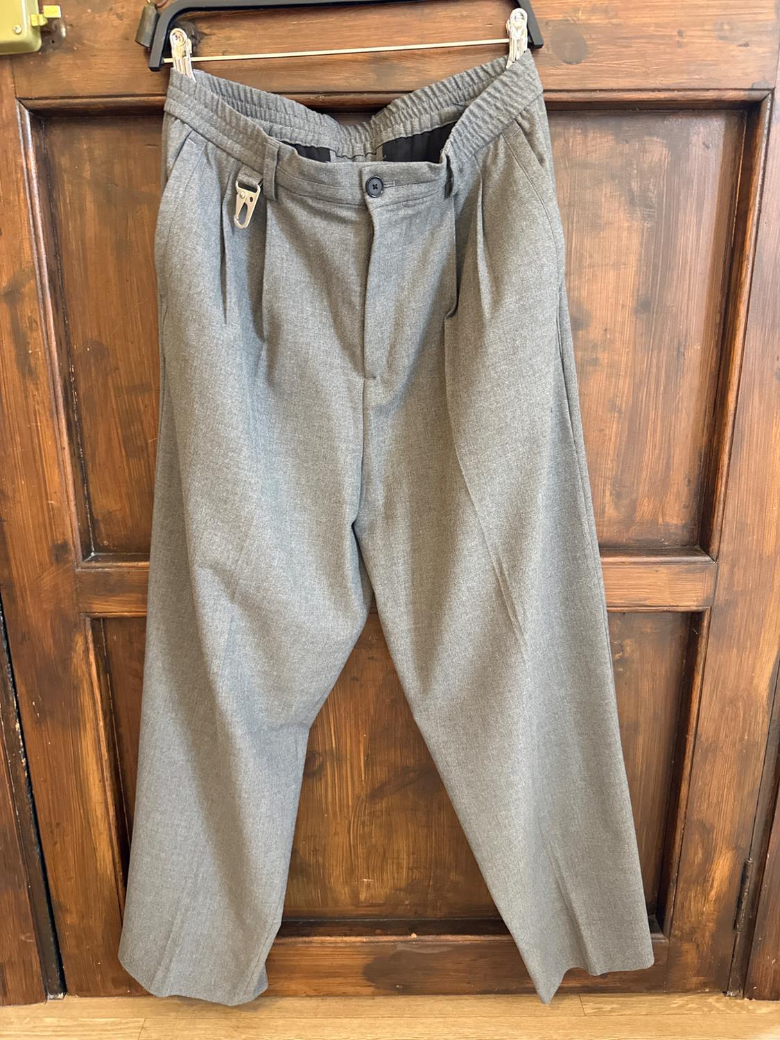 Grey fabric trousers hanging on a hanger over a wooden door. Pleated design with an elasticated waist.