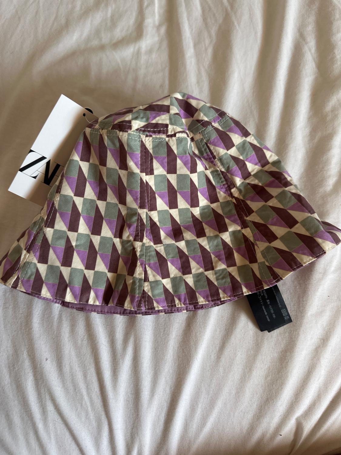 Bucket hat with geometric print in shades of purple and green on a white bed. Visible label with partial text.