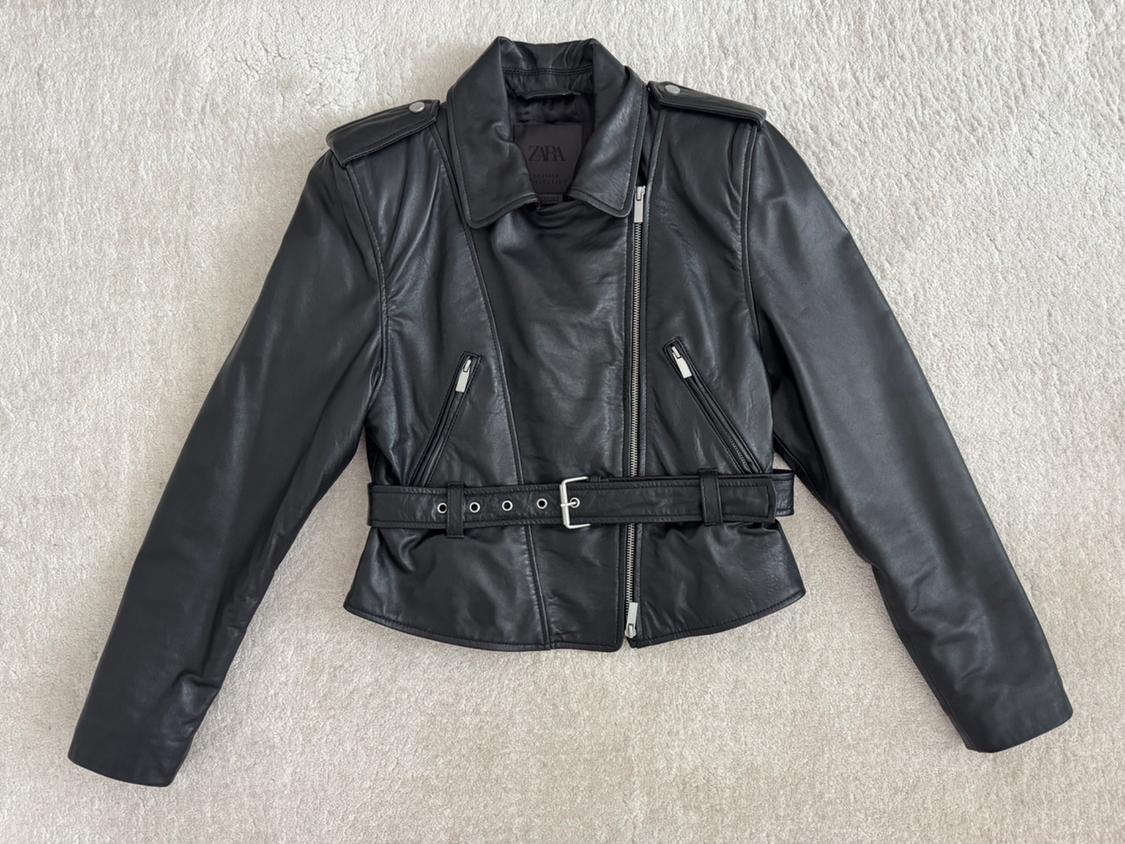 Black faux leather jacket with a diagonal zip, zip pockets, and an adjustable belt on a light background.