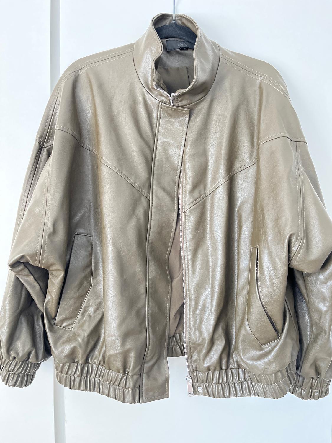 Beige faux leather jacket with front zip, side pockets, and elasticated cuffs, hanging on a hanger.
