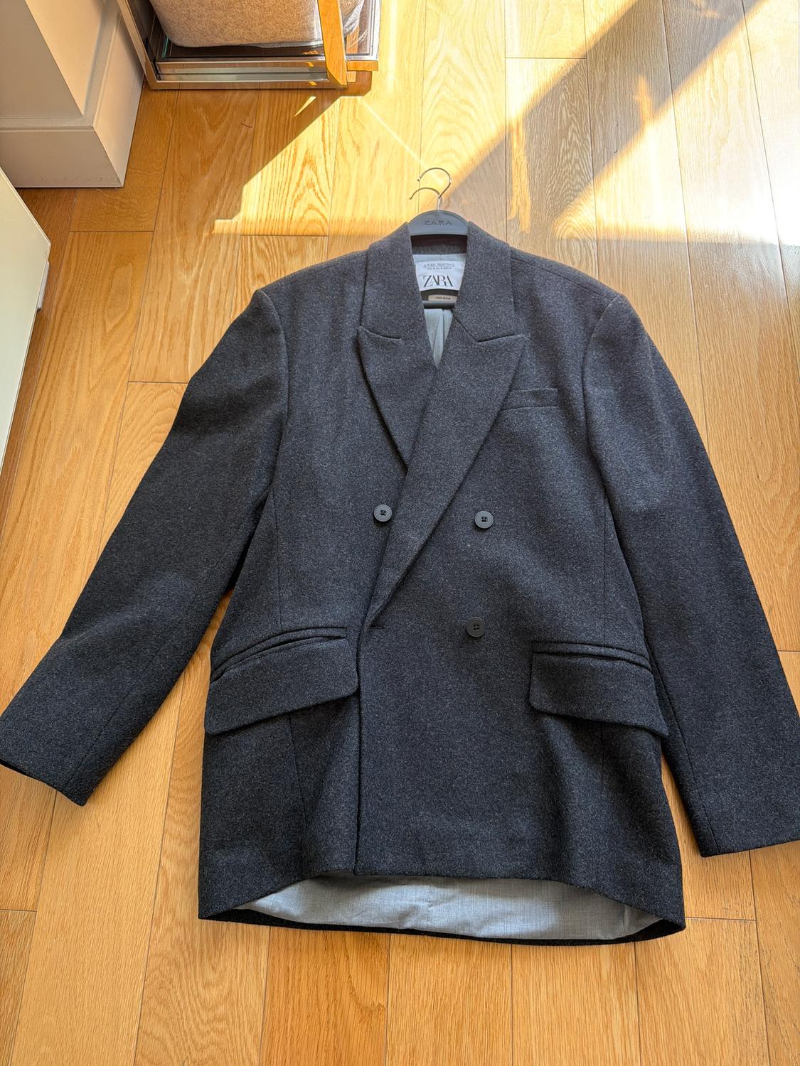 Dark gray double-breasted jacket on light wood floor. Visible tag on hanger with text "ZARA".