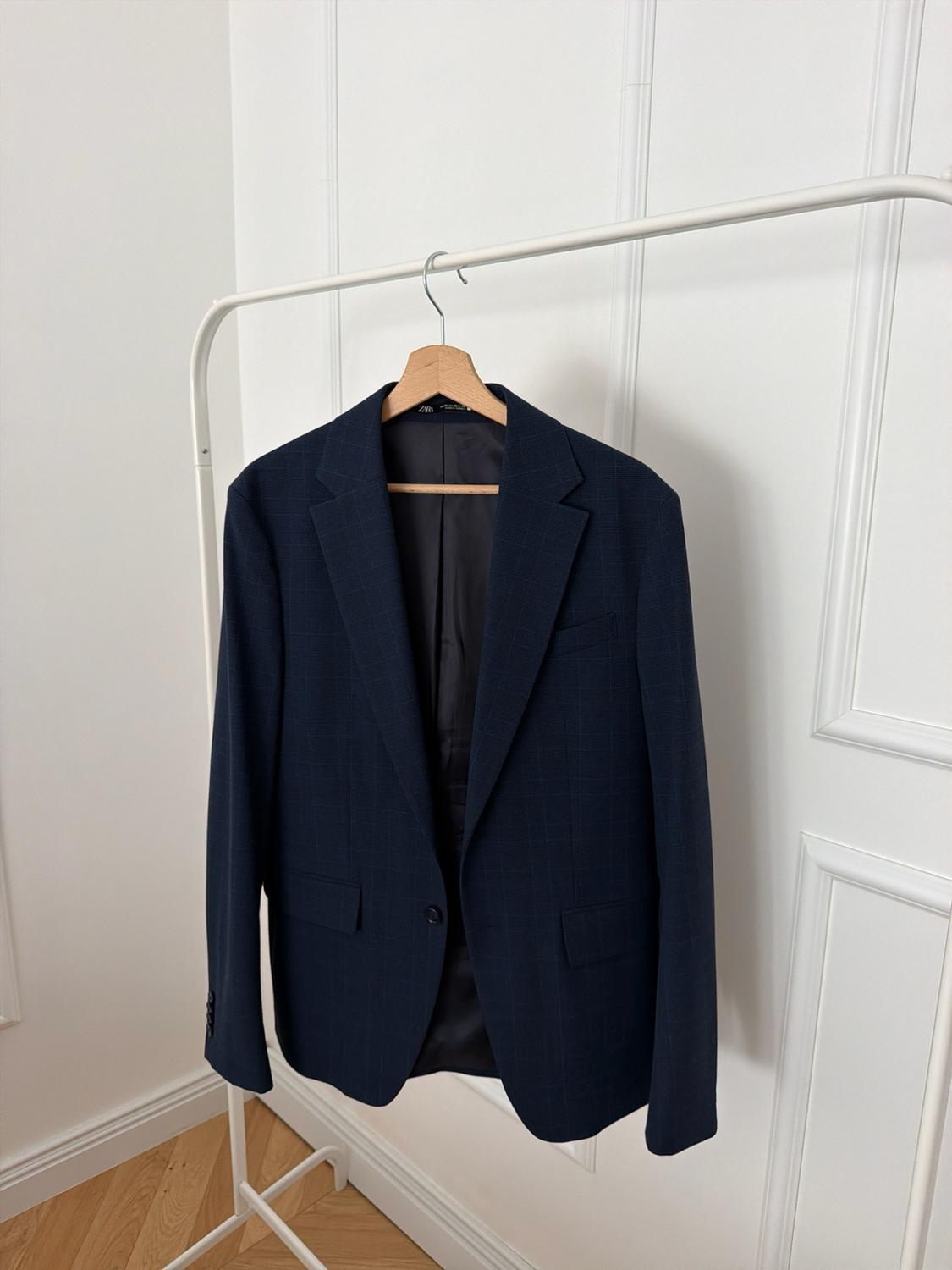 Navy blue blazer with a subtle check pattern on a wooden hanger on a white coat rack. White wall with mouldings in the background.