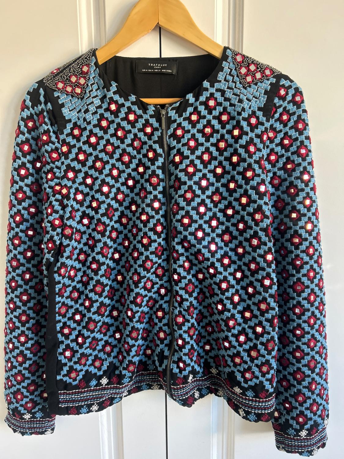 Hanging jacket with a geometric design in blue and red, bead details on the shoulders, black background. "TRAFALUC" label.